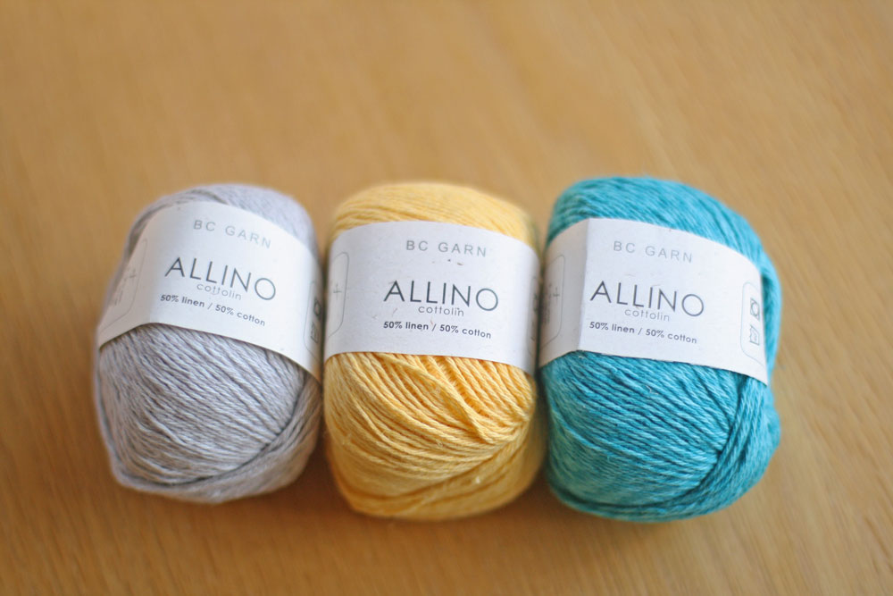 Three skeins of BC Garn Allino yarn in gray, yellow, and blue colors, displayed on a wooden surface.