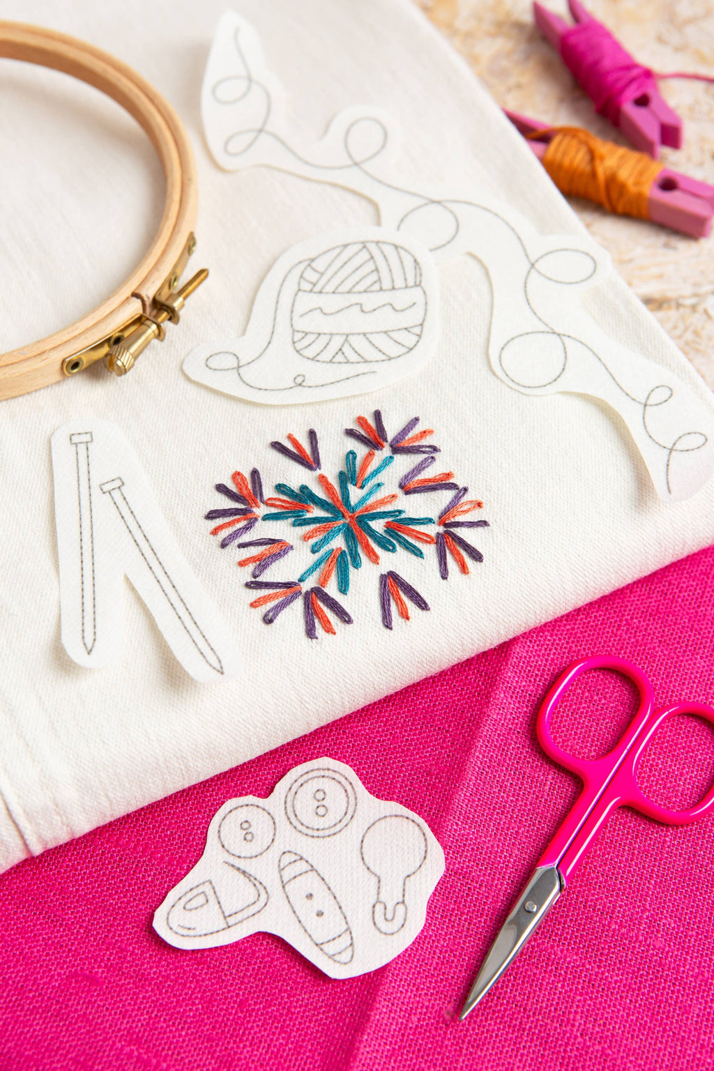 Embroidery hoop, fabric with embroidered design, scissors, and yarn on a pink surface