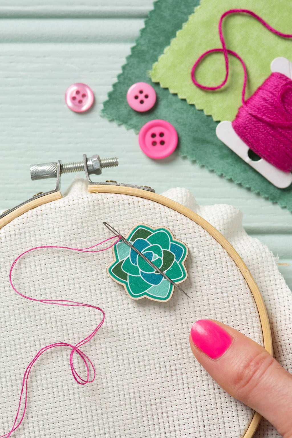 Embroidery hoop with green flower design, pink thread, and buttons on a light green background