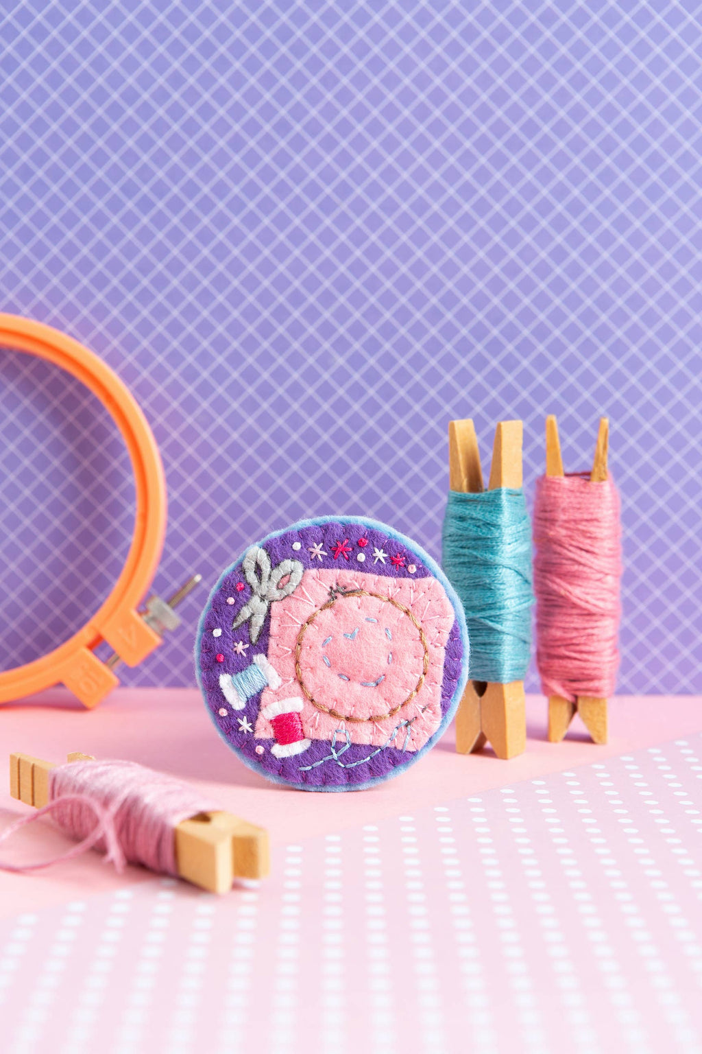 Crafting materials including an embroidery hoop, spools of thread, and a decorative item on a pink and purple checkered background.