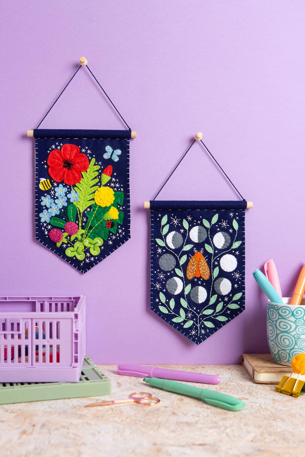 Two decorative wall hangings with floral and moon designs on a purple wall.