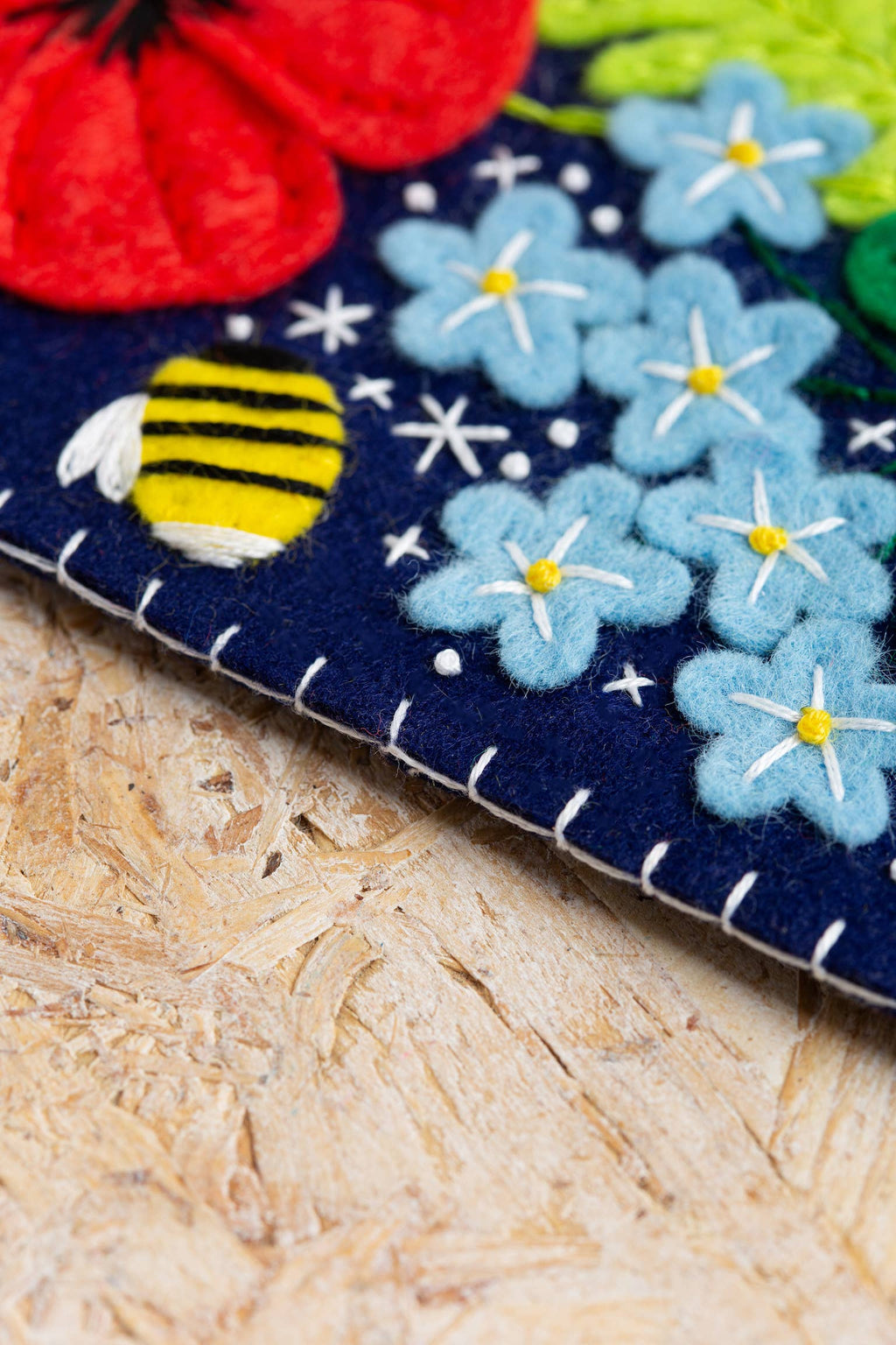 Felt craft with blue flowers and a bee on a dark blue background, placed on a wooden surface.